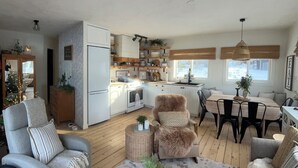 Amplia sala de estar con sofá, sillón y mesa de centro, cocina con armarios blancos y detalles de madera, y comedor con mesa y sillas.