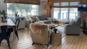 Una sala de estar acogedora con suelo de madera, un sofá gris, un sillón con una manta de piel, una mesa de comedor con sillas y un ventanal con vistas a un lago.