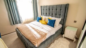 A bedroom with a tufted headboard, a bed with a beige blanket, and a nightstand with a white cabinet. - RoyalGardens Kilimani View Palace Apart  (Nairobi)