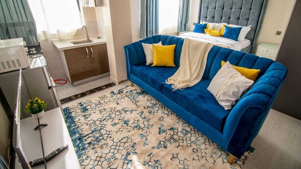 A hotel room with a blue sofa, a bed with a grey headboard, a small desk, and a window with curtains. - RoyalGardens Kilimani View Palace Apart  (Nairobi)