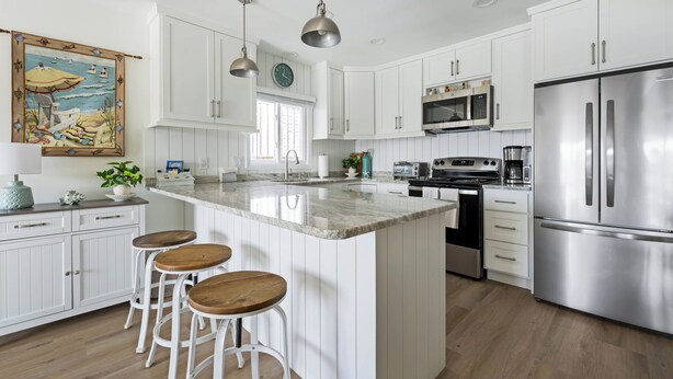 A modern kitchen with white cabinets, stainless steel appliances, a large island with stools, pendant lighting, and a decorative painting.