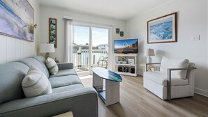 A modern living room with a grey sofa, a white armchair, a coffee table, and a TV on a stand. There is a balcony with a view of the sea.
