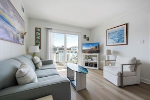 Een moderne woonkamer met een grijze bank, een witte fauteuil, een salontafel en een televisie op een meubel. Er is een balkon met uitzicht op zee.
