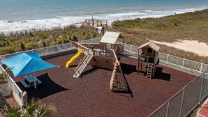 A playground with a slide and climbing structures, surrounded by a fence, overlooking the ocean.