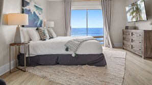 Una camera da letto con un letto, un ventilatore a soffitto, un comodino con lampada, una cassettiera e un'ampia finestra vista mare.