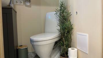 A bathroom with a toilet, a trash bin, a potted plant, and a roll of toilet paper.