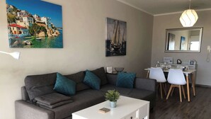 Una sala moderna con un sofá gris, cojines verde azulado, una mesa de centro blanca y un comedor con sillas blancas y una mesa de madera.