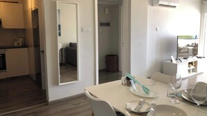Una cocina moderna con armarios blancos, un comedor con una mesa blanca dispuesta para dos personas, y un espejo grande en la pared.