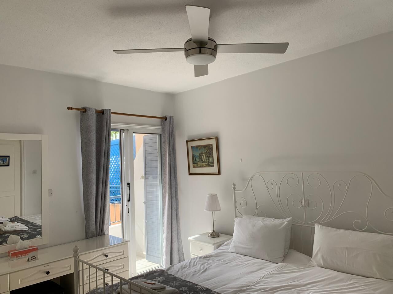 A bedroom with a bed, a nightstand, a lamp, a ceiling fan, a window with curtains, and a door leading to an outdoor area.