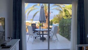 Ein Zimmer mit einer Glastür, die auf einen Sitzbereich im Freien mit Tisch und Stühlen führt. Blick auf den Strand, auf dem Tisch steht eine kleine Statue.