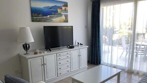 Ein Wohnzimmer mit Fernseher auf einer Kommode, einer Couch, einem Tisch und Blick ins Freie durch ein Fenster.