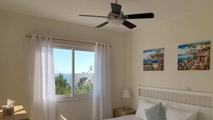Ein Schlafzimmer mit Bett, Nachttisch, Fenster mit Vorhängen, Deckenventilator und zwei Wandbildern.