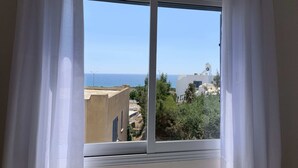 Ein Fenster mit weißen Vorhängen, freier Blick auf das Meer und Gebäude im Hintergrund.