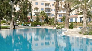 Una piscina de borde curvo, rodeada de palmeras y un edificio con balcones al fondo.