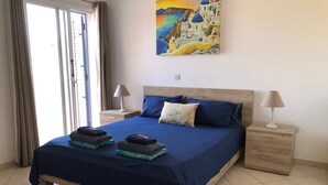 Une chambre à coucher avec un grand lit, deux tables de chevet, un tableau représentant un paysage côtier et une porte-fenêtre coulissante.
