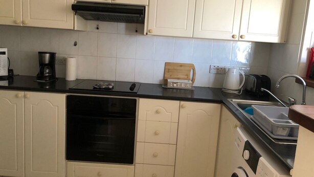 Una cocina compacta con armarios blancos, encimera negra, horno empotrado, cafetera, tostadora y pileta.
