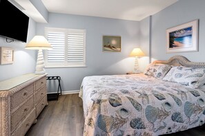 Une chambre à coucher avec un lit, deux lampes de chevet, une télévision, une commode en osier, une fenêtre avec des volets et deux tableaux encadrés accrochés au mur.