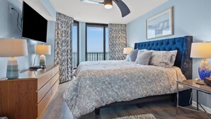 Ein Schlafzimmer mit einem großen Bett, einem an der Wand montierten Fernseher, einem Schminktisch und einem Fenster mit Vorhängen.