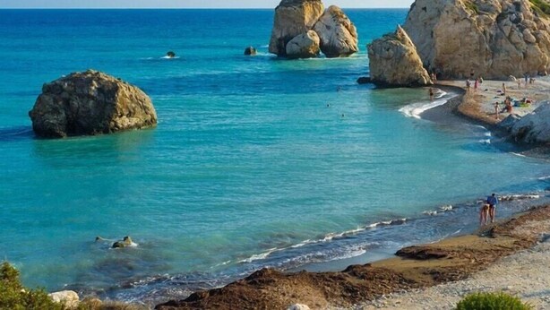 A coastal scene with clear turquoise waters, rocky formations, and a sandy beach with people. - Kings Palace - Modern Penthouse | Incredible Sea Views | Pool (Paphos)