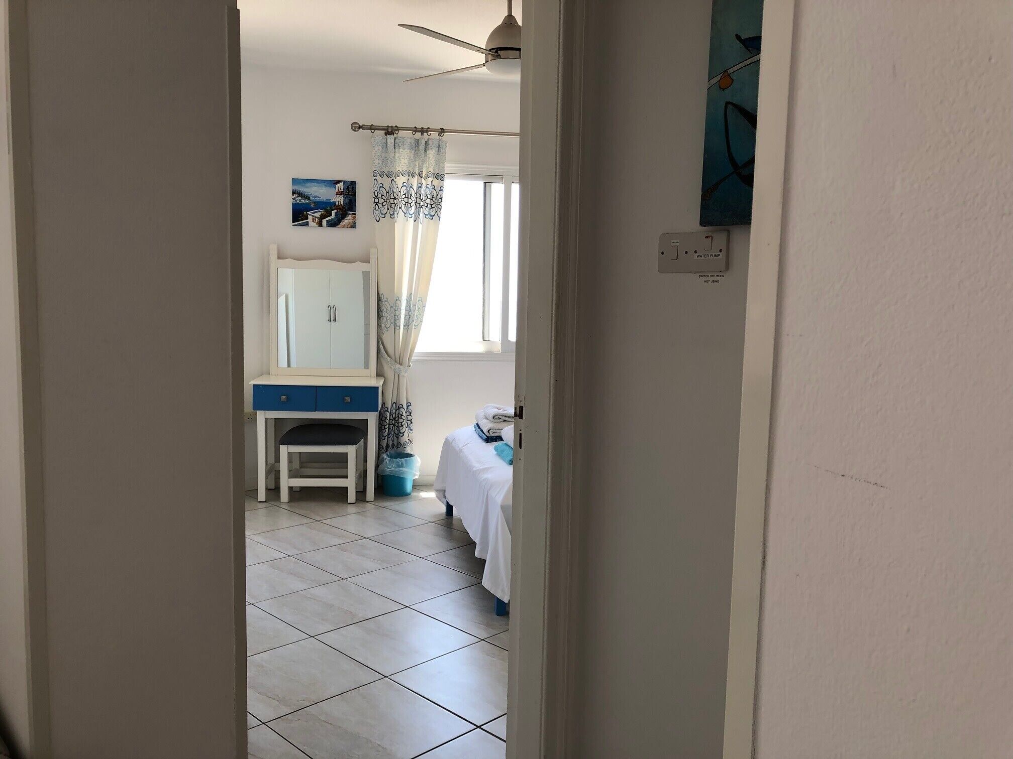 A narrow hallway leading to a bedroom with a bed, a chair, a desk with a mirror, and a window with curtains.