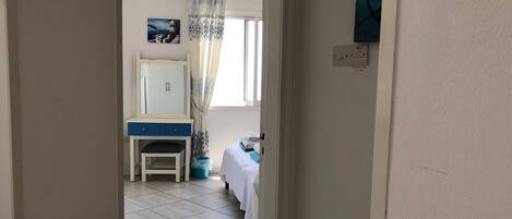 A narrow hallway leading to a bedroom with a bed, a chair, a desk with a mirror, and a window with curtains.