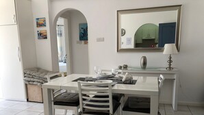 A dining area with a white table and chairs, a mirror, and a lamp. There are paintings on the wall and a view of the outside. - Kings Palace - Modern Penthouse | Incredible Sea Views | Pool (Paphos)