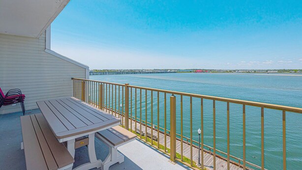 A balcony with a wooden table and chairs, overlooking a calm body of water and a distant pier. - Beautiful Bayfront Townhouse located downtown Ocean City. Free WiFi, Elevator (Ocean City)