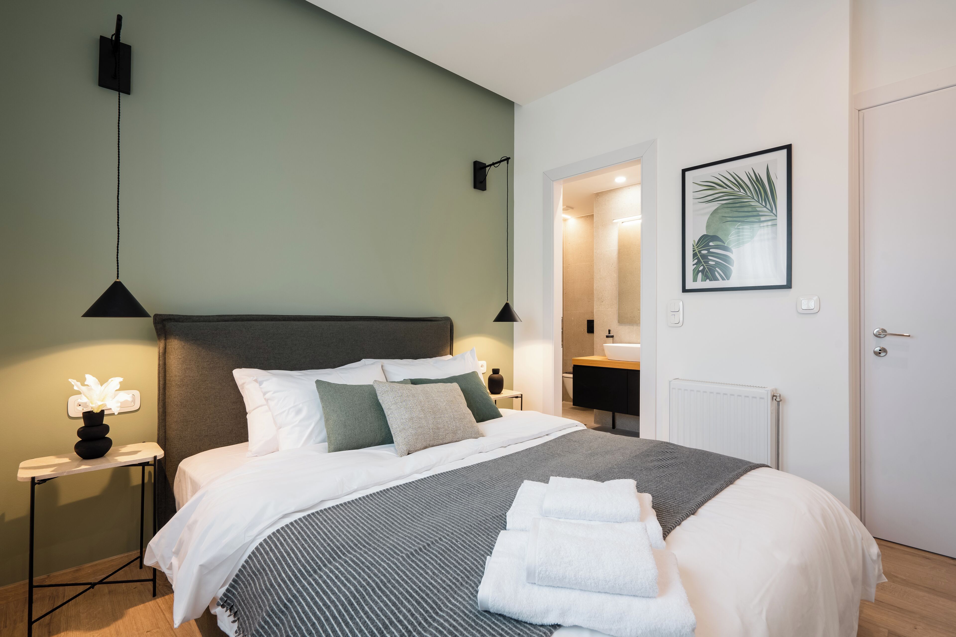 A neatly made bed with white linens, a grey striped blanket, and a stack of white towels. There is a small bedside table with a lamp and a vase of flowers. The room has a green wall, a framed plant on the wall, and a door leading to a bathroom.