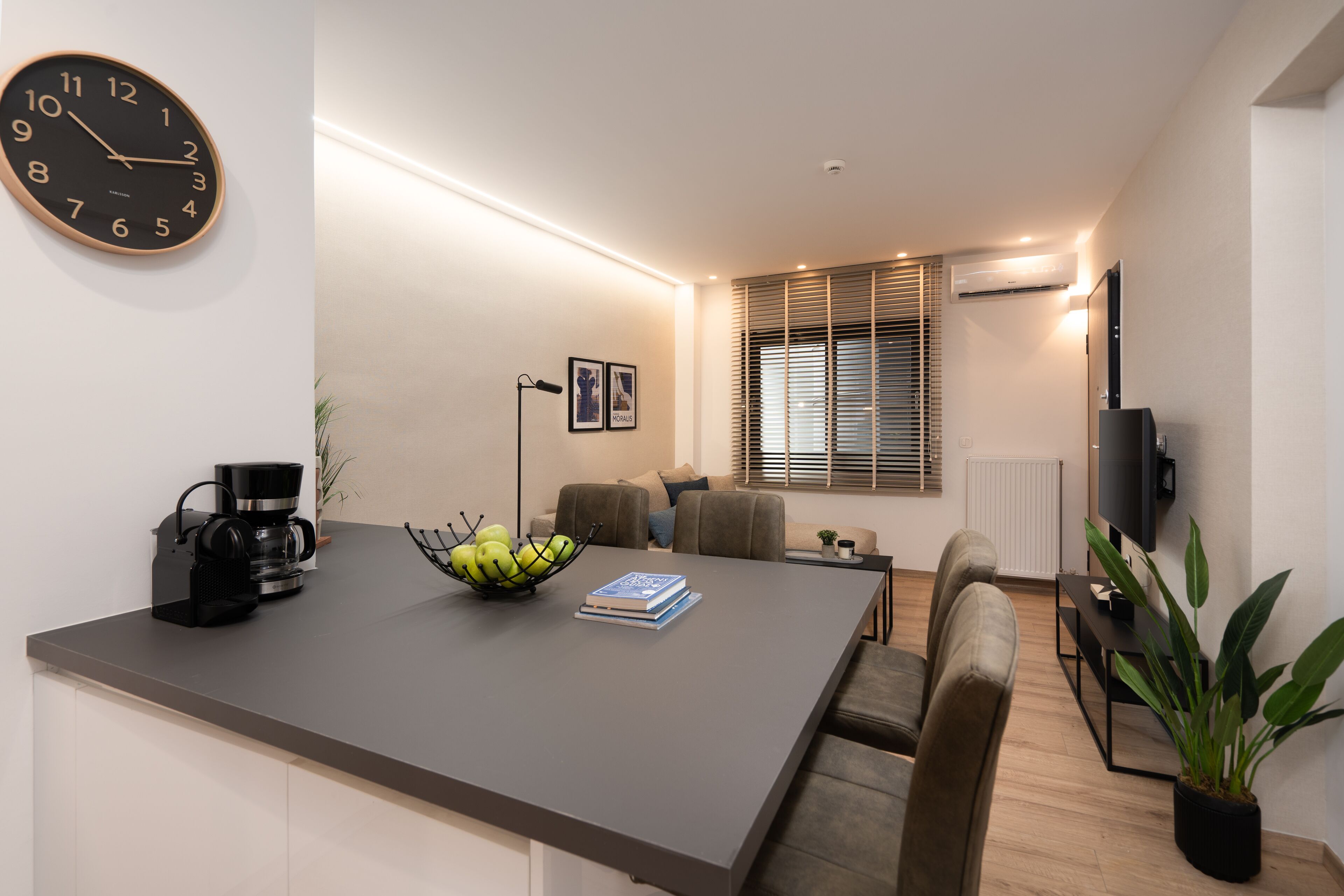 A modern living room with a dining area, a coffee machine, a bowl of fruit, and a flat-screen TV.