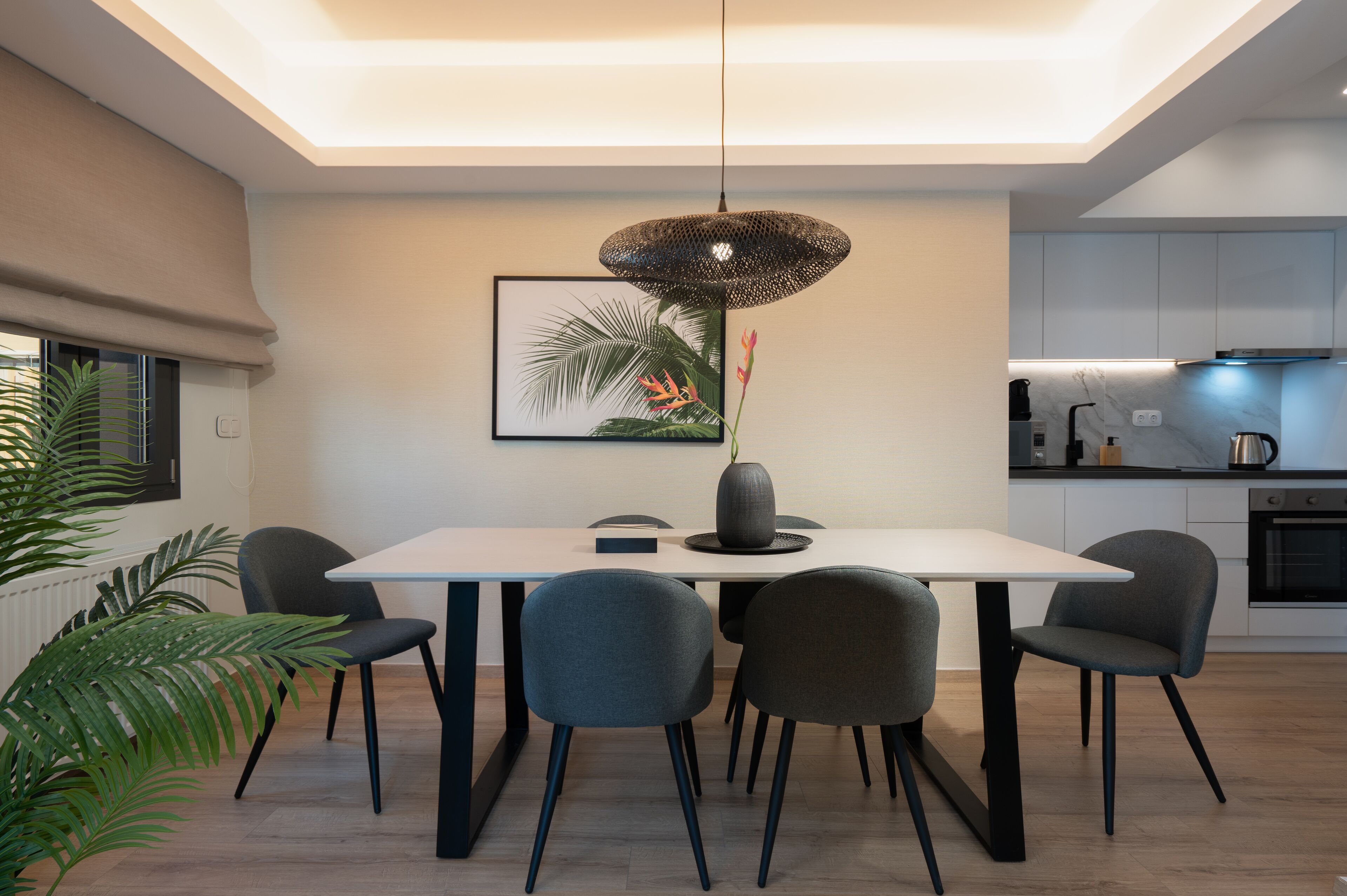 Een moderne eetruimte met een witte tafel, vier grijze stoelen, een zwarte hanglamp en een lijst met een afbeelding van een tropisch landschap aan de muur.