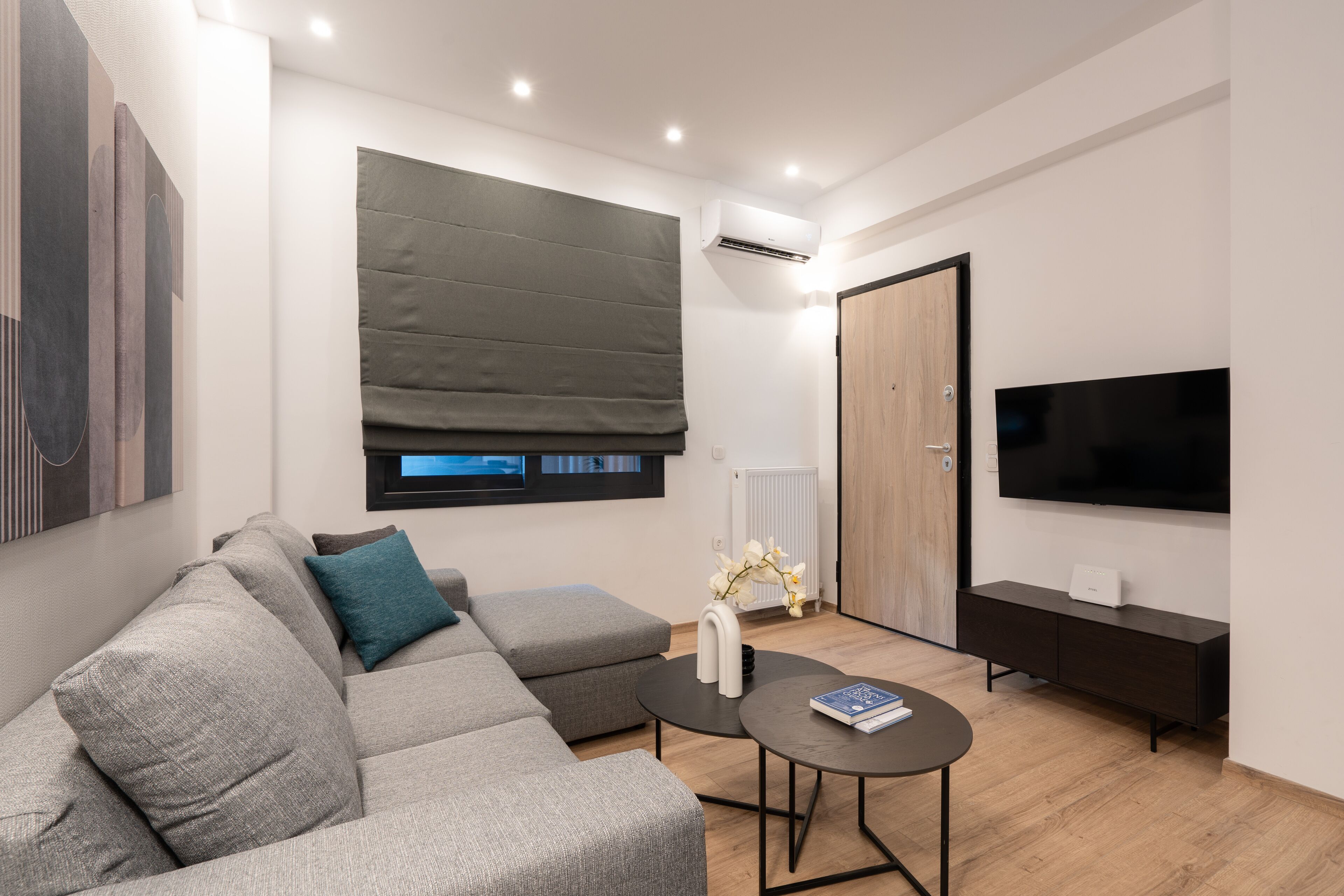 A modern living room with a grey sectional sofa, a small round coffee table, a dark wooden TV stand, and a large window with a green blind.