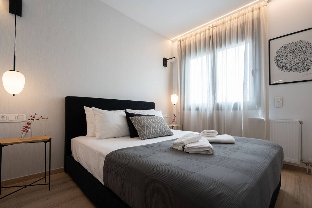 Une chambre à coucher moderne avec un grand lit, des tables de chevet, une fenêtre avec des rideaux et un luminaire fixé au mur.
