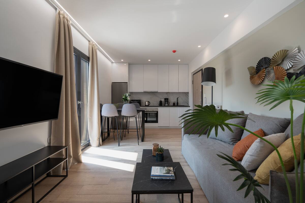 Un salon moderne avec un téléviseur à écran plat, un canapé gris, une table basse et un coin repas avec des chaises, et une cuisine en arrière-plan.