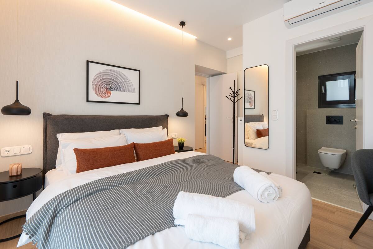 Une chambre d’hôtel moderne dotée d’un grand lit, de tables de chevet, d’un téléviseur à écran plat et d’une salle de bain visible à travers une porte ouverte.