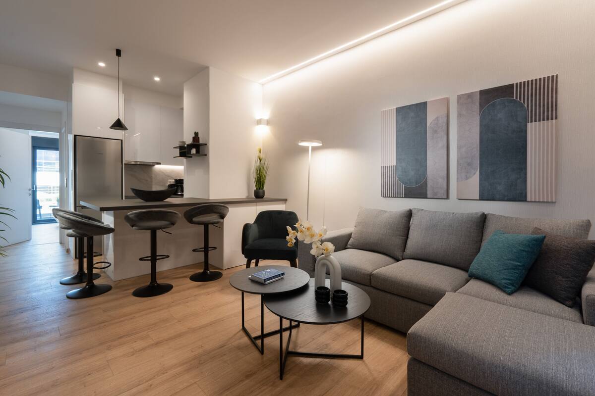 Un salon moderne avec un canapé gris, une table basse ronde et un tabouret de bar noir. On y trouve également un coin cuisine avec une chaise noire et une plante.