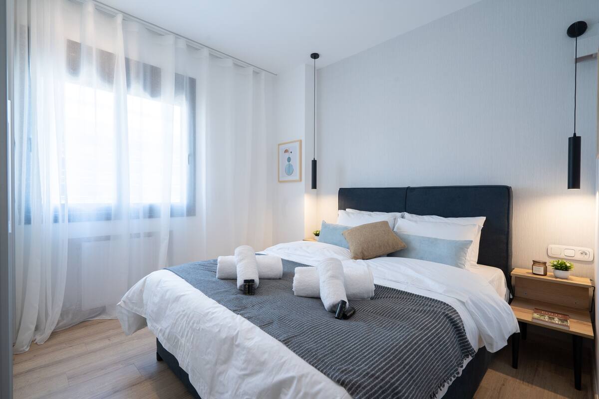 Un lit bien fait, avec une literie blanche et grise, une table de chevet avec une lampe et une fenêtre ornée de rideaux légers.