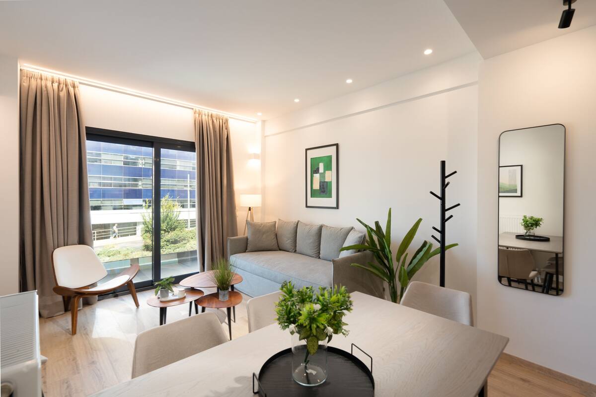 Une chambre d’hôtel moderne avec un canapé, une table à manger et des chaises, une grande fenêtre avec des rideaux, une plante en pot et un miroir au mur.