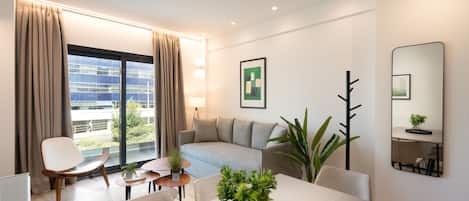 A modern hotel room with a sofa, dining table, and chairs, a large window with curtains, a potted plant, and a mirror on the wall.