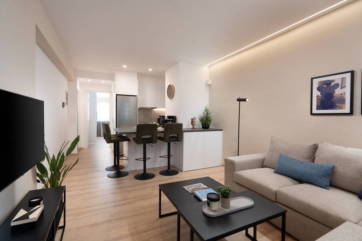 Un salon moderne avec un canapé, une table basse et un téléviseur. Il y a également un coin cuisine avec des tabourets de bar et une table à manger.