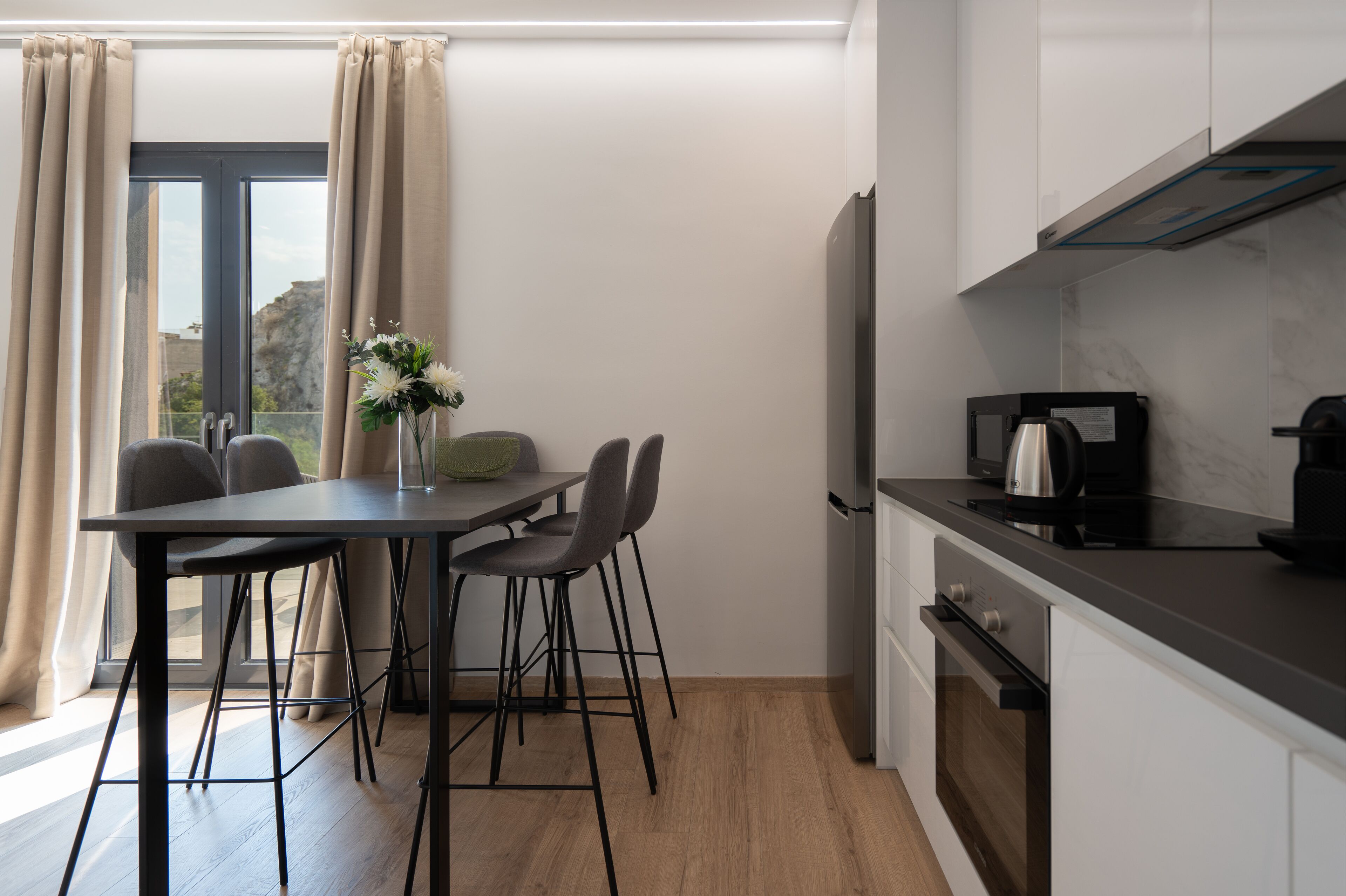 A modern kitchen with a dining area, including a table with chairs, a vase with flowers, and a coffee maker.