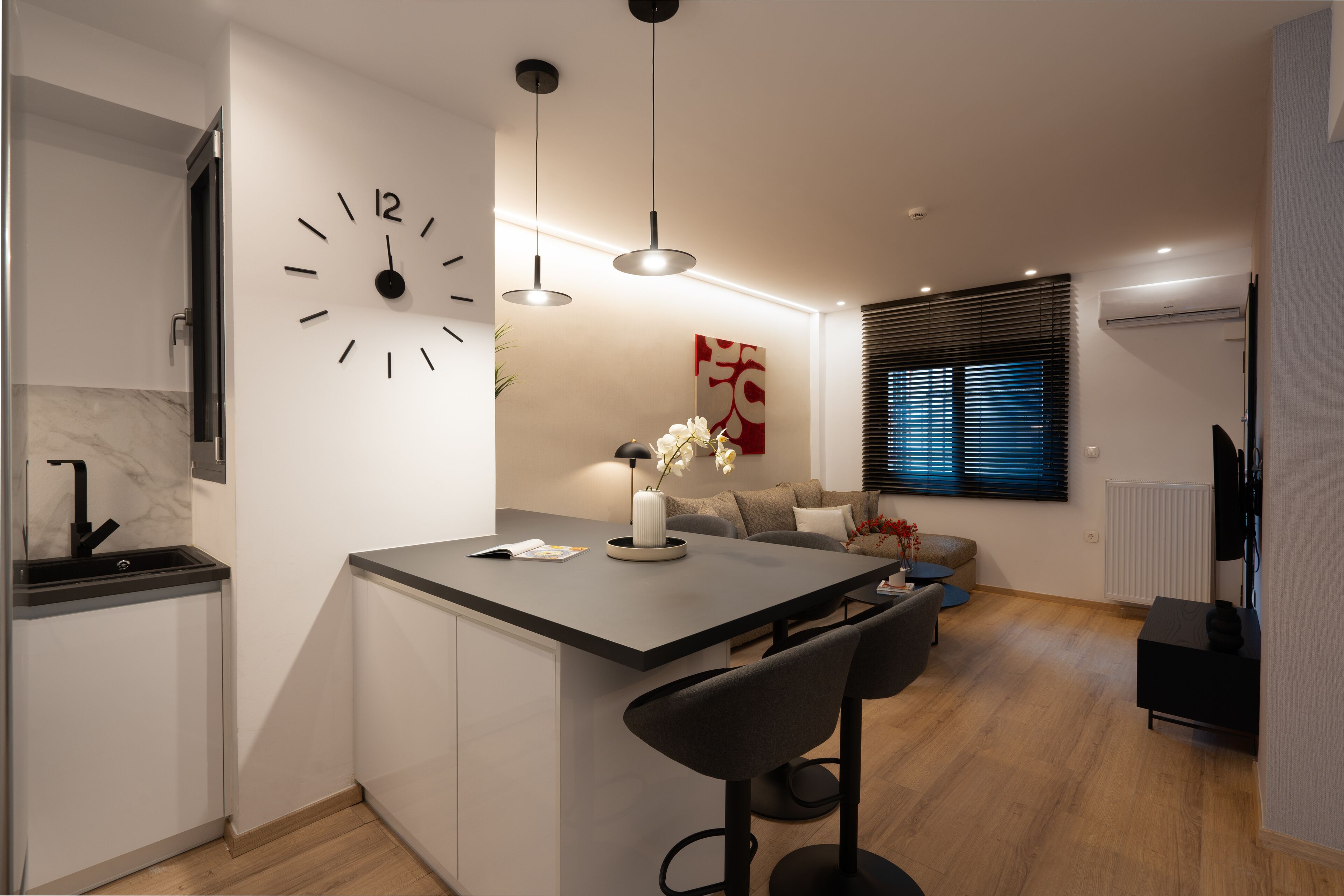 A modern kitchen with a central island, bar stools, a clock, pendant lights, and a dining area with a sofa and a coffee table.