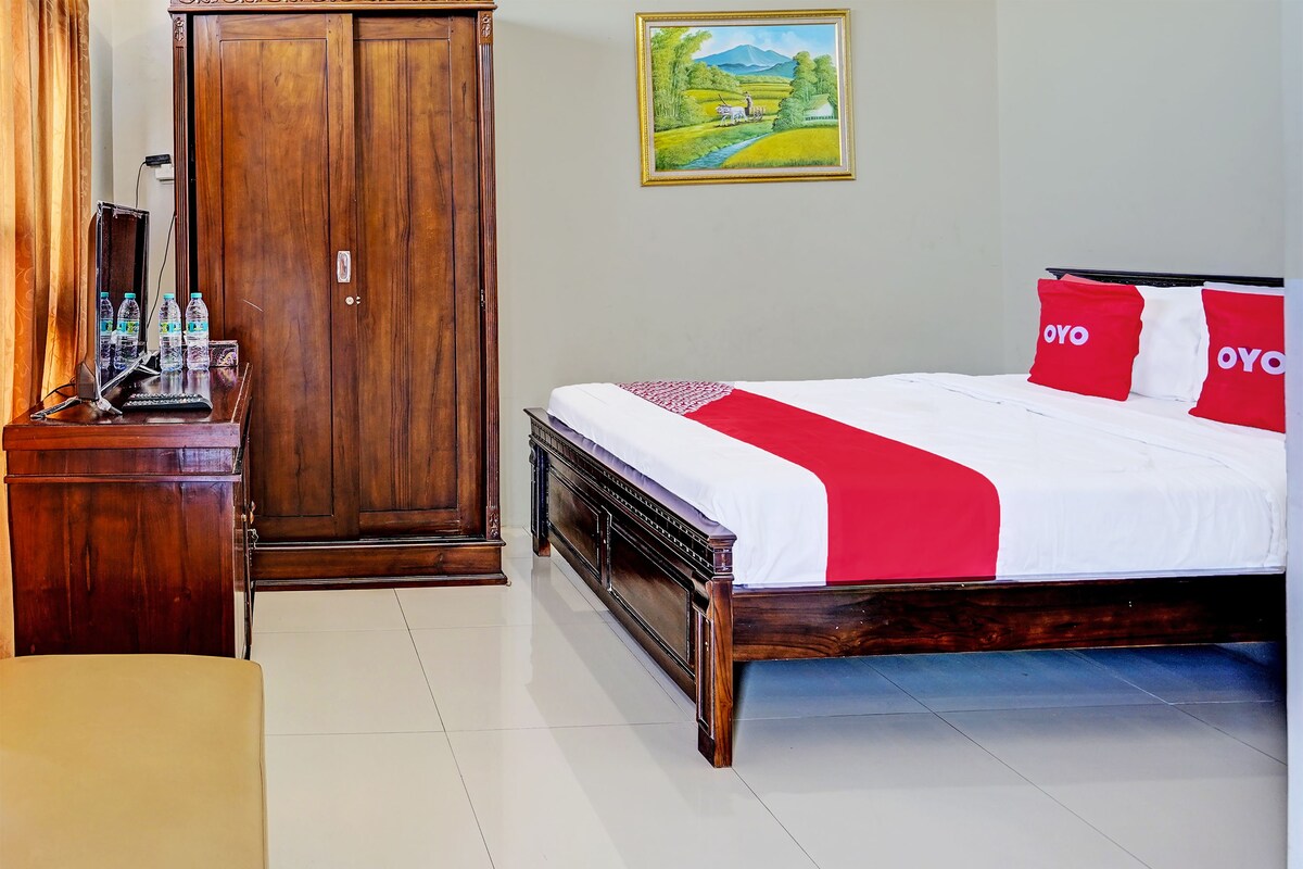 A hotel room with a wooden wardrobe, a desk with a computer and bottles, a bed with red and white bedding, and a painting on the wall.