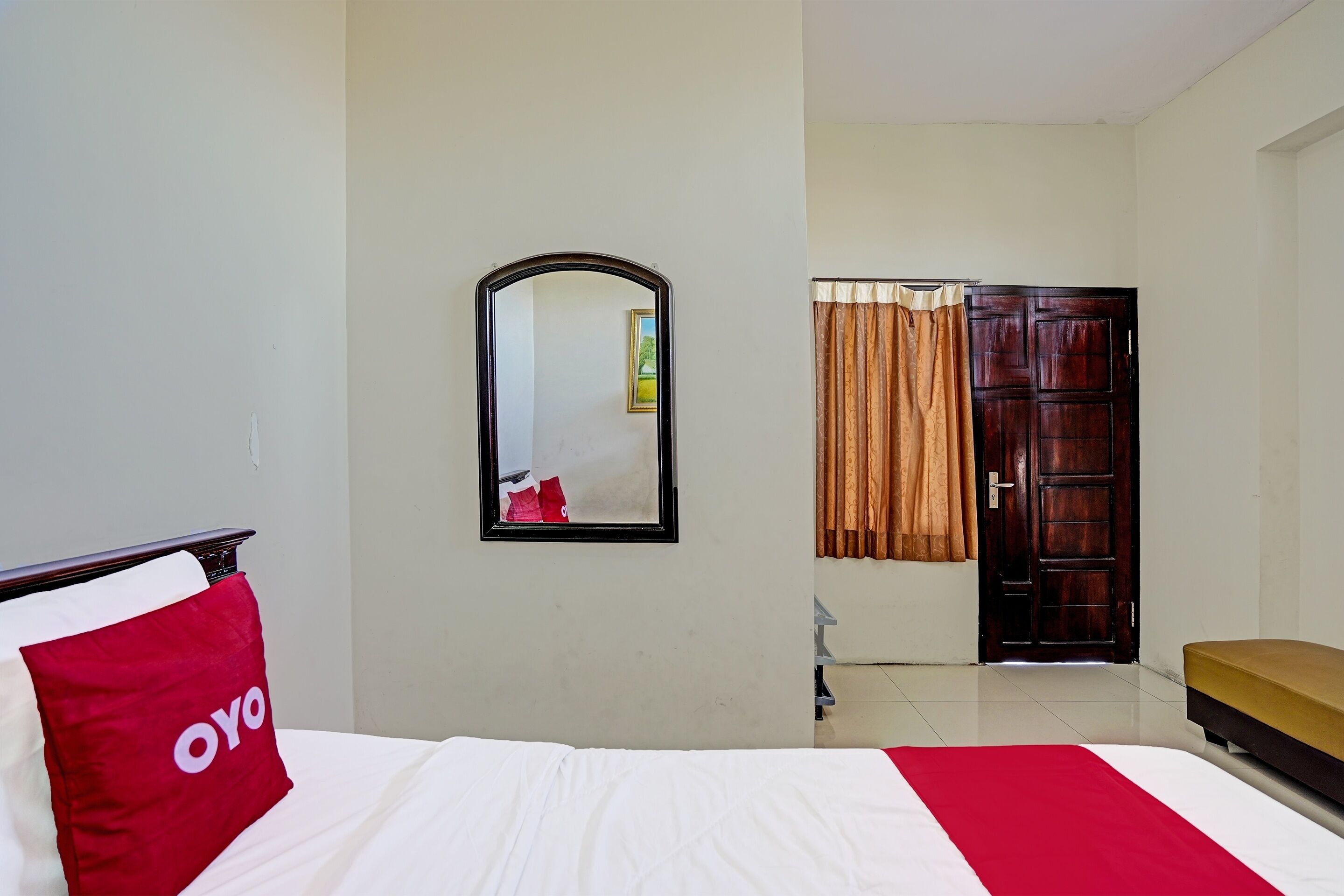 A hotel room with a bed, a red pillow, a mirror, a wooden door, and a bench.