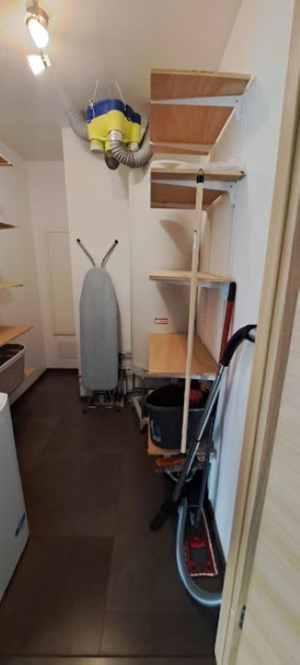 A narrow kitchen with wooden shelves, a drying rack, and a vacuum cleaner.