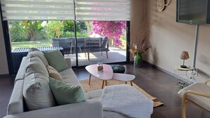 Un salon moderne comprenant un canapé, une petite table basse, une table d’appoint et un fauteuil inclinable. Une grande fenêtre offre une vue sur un jardin et une terrasse à l’extérieur.