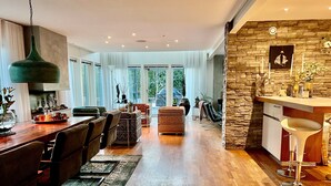Amplia sala de estar con piso de madera, una mesa de comedor con sillas, un sofá y una zona de cocina con pared de piedra y taburetes de bar.