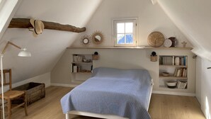 A cozy attic bedroom with a sloped ceiling, a bed with a blue quilt, wooden shelves with books and decorative items, a small window, and a rustic hat hanging on the wall. - Le Vieux Logis, an idyllic setting (Varengeville-sur-Mer)