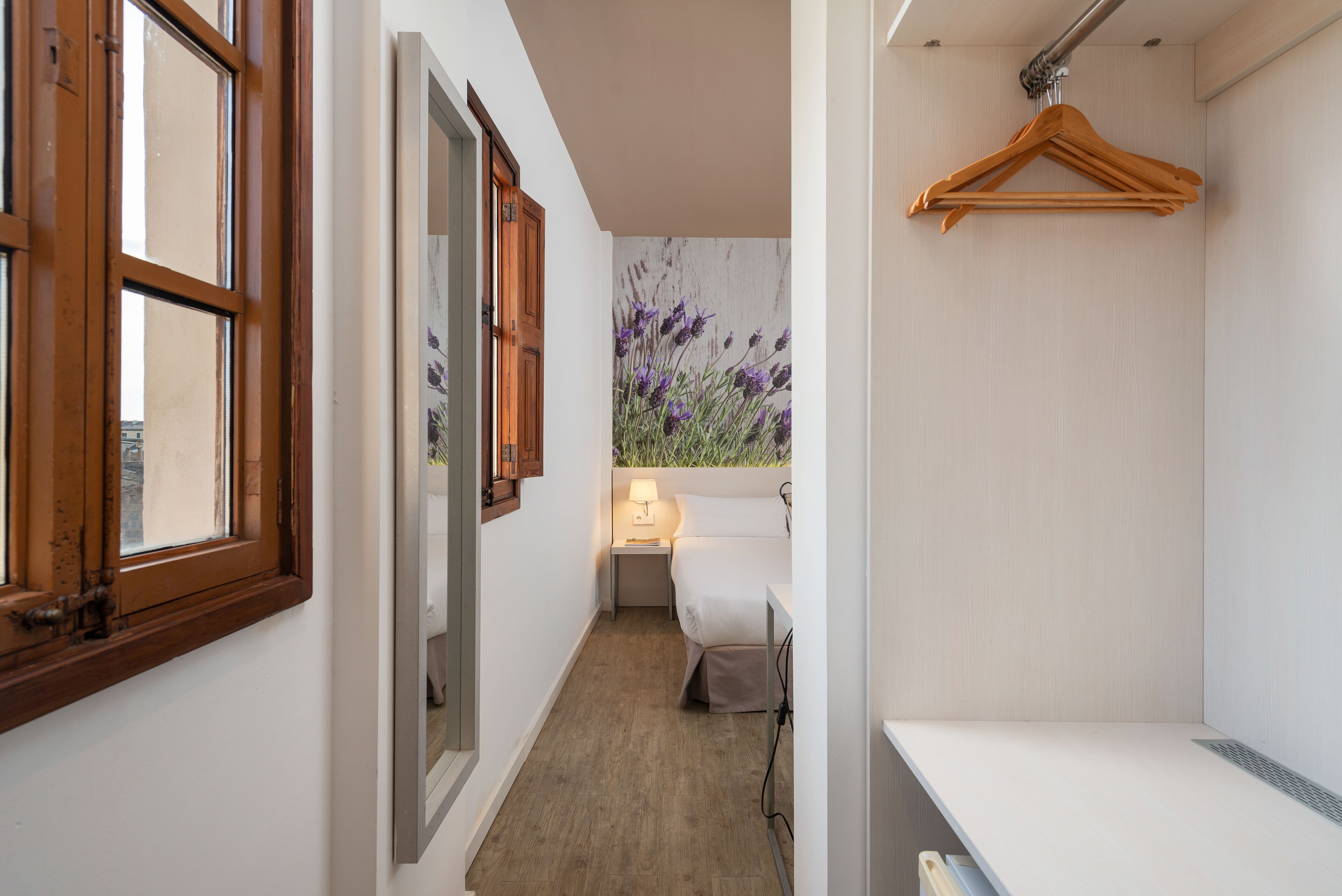 Un pasillo estrecho con una puerta de madera, una mesita de noche pequeña con una lámpara y una cama con edredón blanco.