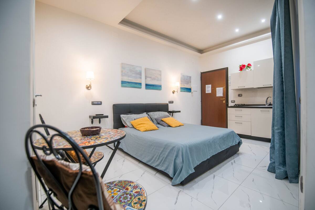 A bedroom with a bed, a small table, and a chair. There is a kitchenette in the background.