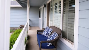 Eine überdachte Veranda mit einem Korbstuhl und Kissen, einem weißen Geländer und einem Fenster mit Jalousien.